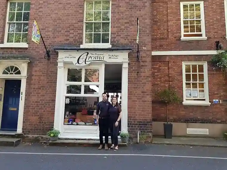 Alt text: A brick building with a storefront labeled "Aroma". Two people, a man and a woman, are standing in the doorway wearing dark shirts. The building has multiple windows and a blue door to the left of the storefront. There are small plants in pots near the entrance and a colorful flag on display.best cafes in Los Angeles