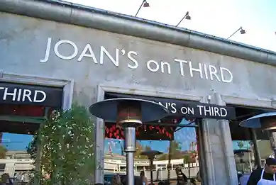 Alt text: Exterior view of a restaurant called "Joan's on Third". The storefront features large windows and a gray awning. The restaurant name is displayed in large white letters above the entrance. Outdoor seating is visible, and there are string lights and plants decorating the exterior.