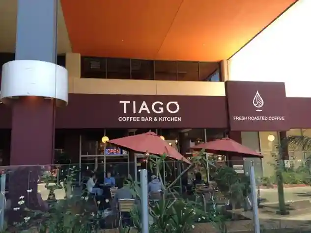 Alt text: Exterior of Tiago Coffee Bar & Kitchen. The storefront has a dark awning with the business name in white letters. To the right, there's a sign indicating "Fresh Roasted Coffee". Outdoor seating area visible with pink umbrellas and patrons seated at tables. The building has a modern design with an orange-lit overhang.