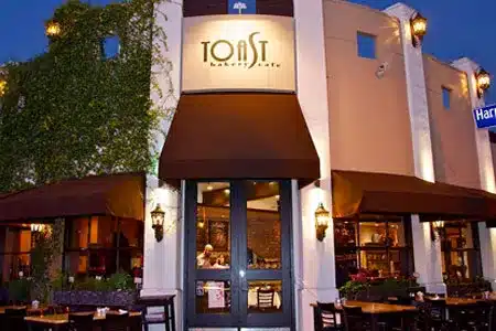 Alt text: Exterior of Toast Bakery Cafe at night. The building has a curved facade with the restaurant name illuminated above the entrance. Brown awnings extend over the windows, and outdoor seating is visible. Potted plants and wall-mounted lights decorate the exterior. The warm lighting from inside creates an inviting atmosphere.best cafes in Los Angeles